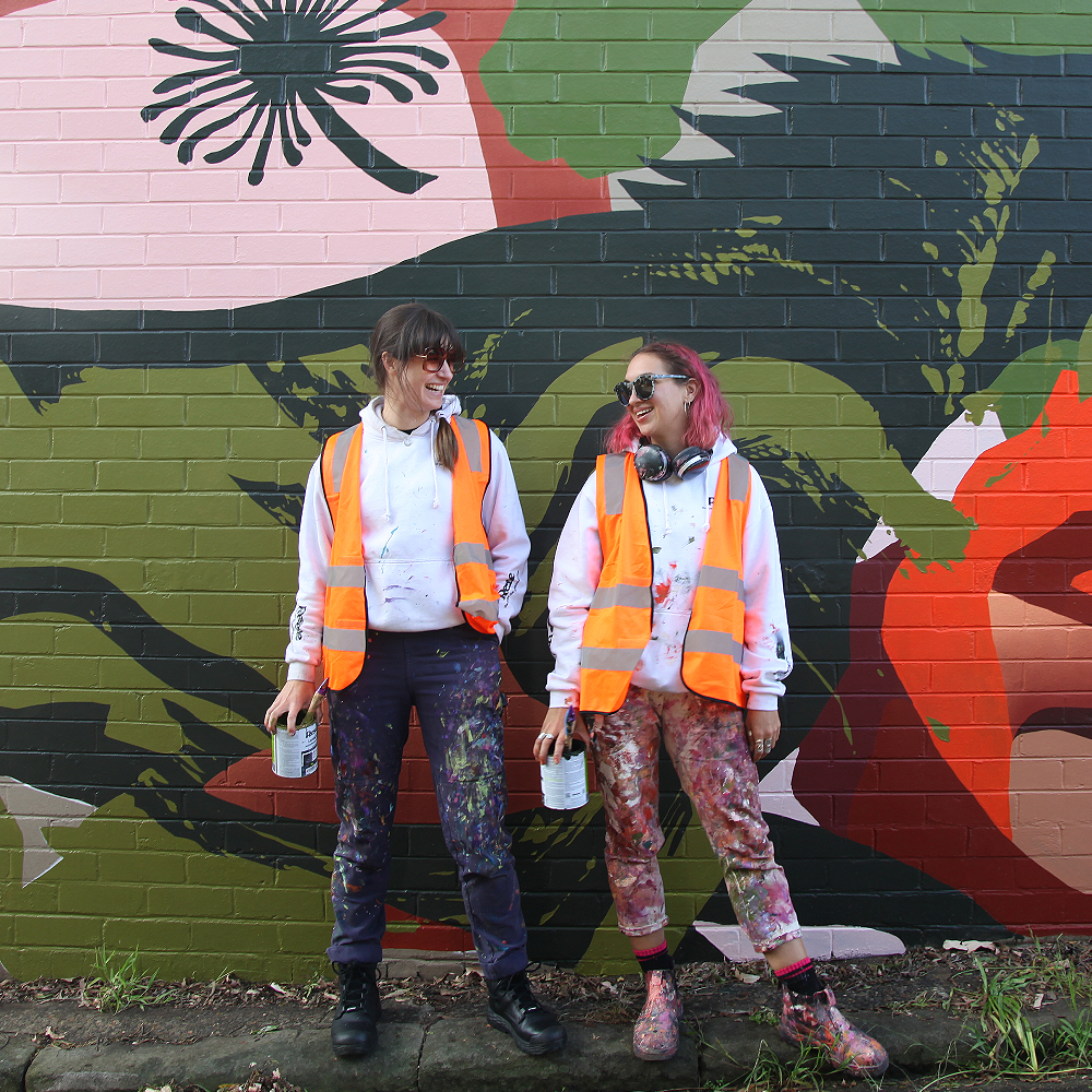 Two artists stand on a small kerb on the street in front of a brick wall adorned with an artwork.
