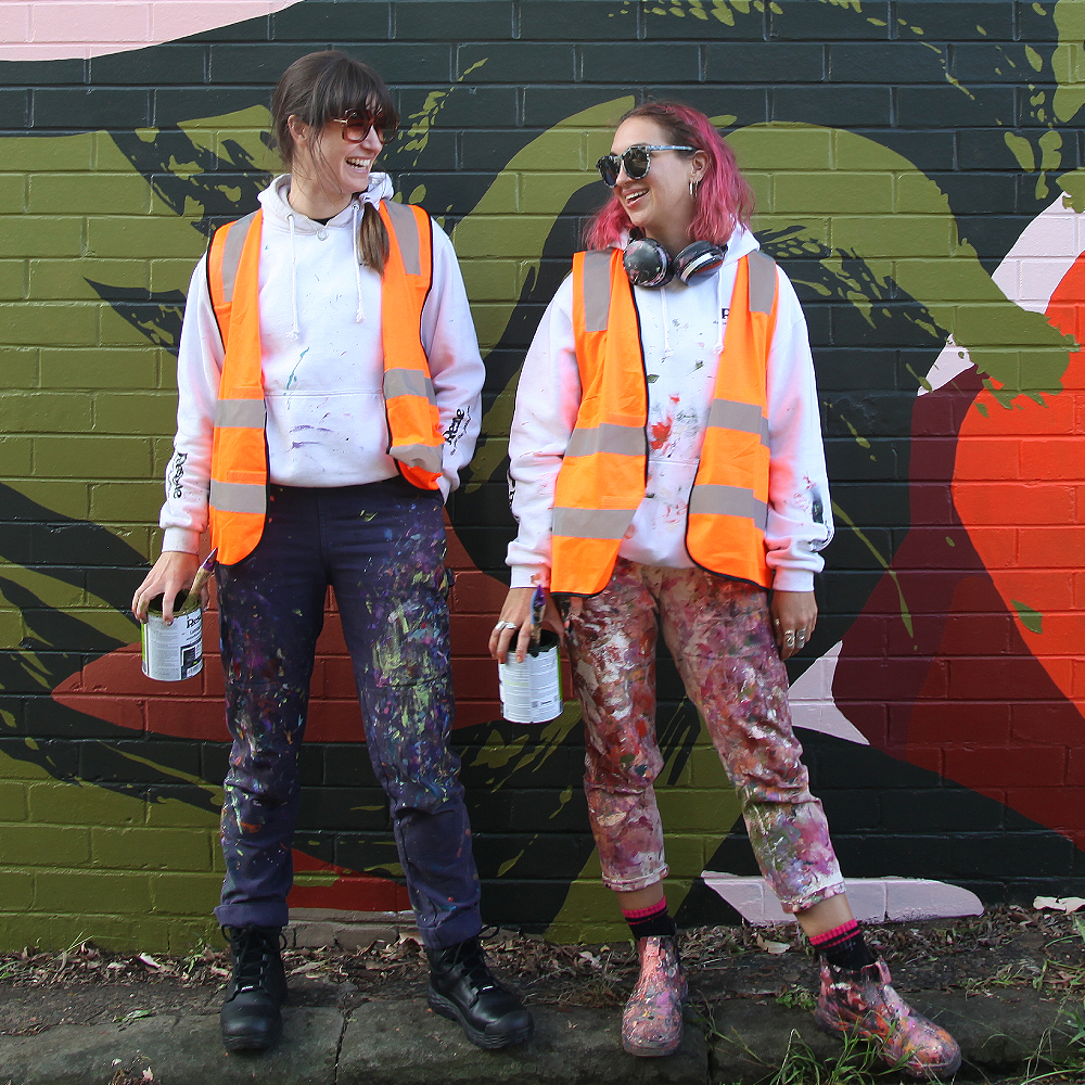  Two artists stand on a small kerb on the street in front of a brick wall adorned with an artwork.