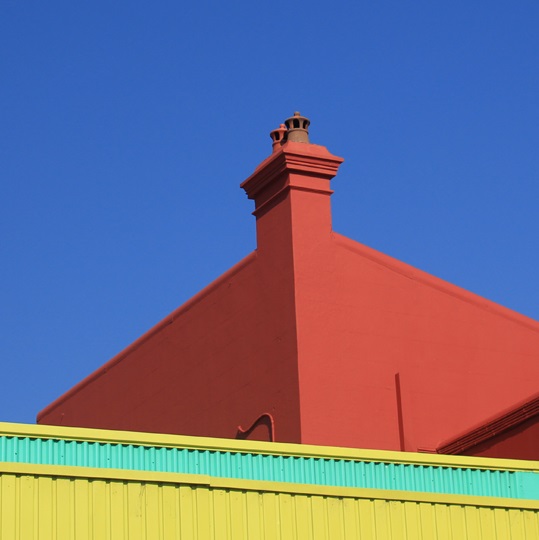 Abstract layered photograph of a bright yellow green and corrugated building, in front of a rendered red building, in front of a plain blue sky