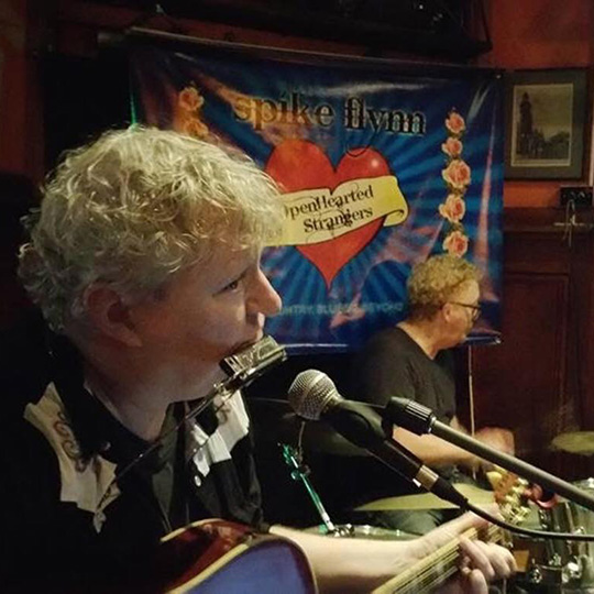 A blonde-haired guitar player with a harmonica and a drummer in an intimate music venue