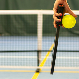 Close up of someone holding a racquet and yellow ball in front of a sporting net