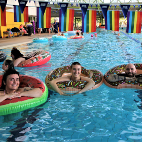 Group of smiling people in an indoor pool on flotation devices. A row of rainbow flags is above.