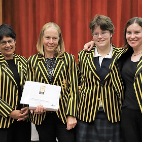 four women standing together wearing black and gold blazers while one holds up an award