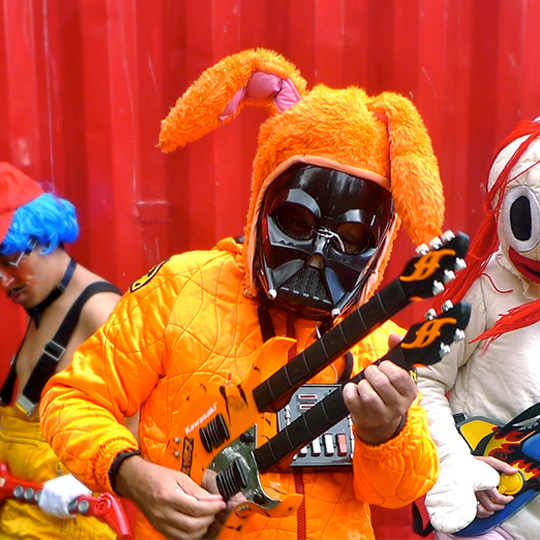 A person in a plush bunny outfit with a Darth Vader mask and chest panel, playing a double-necked toy guitar