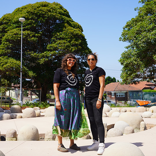 Two women smiling and standing between spiralling rows of stones arranged into an artwork within a park.