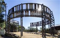 Large circular site specific artwork with tin panels slightly rusted and letters laser cut into the panels sitting on rock platform surrounded by trees and blue sky backdrop