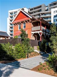 Heritage red brick building with red and creme features with brown fence and natural surrounding with towering white and grey residential buildings in background