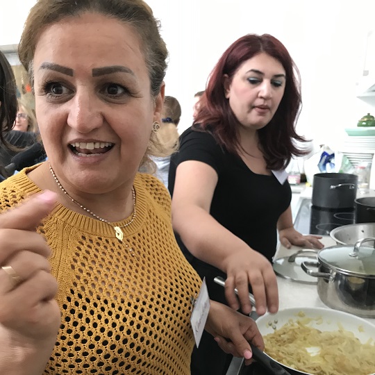 Photo of Wasan in black t-shirt in background, another project participant is in forefront of photo, photo is in a kitchen setting and women are cooking