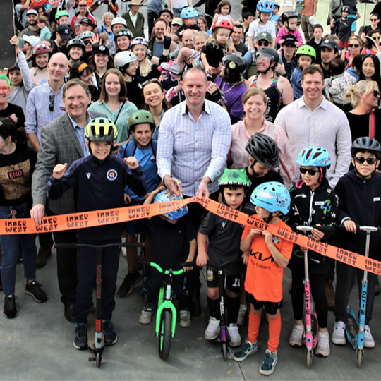 Large crowd of adults and kids, some on bikes and skate boards, behind and orange ribbon being cut by a smiling man 