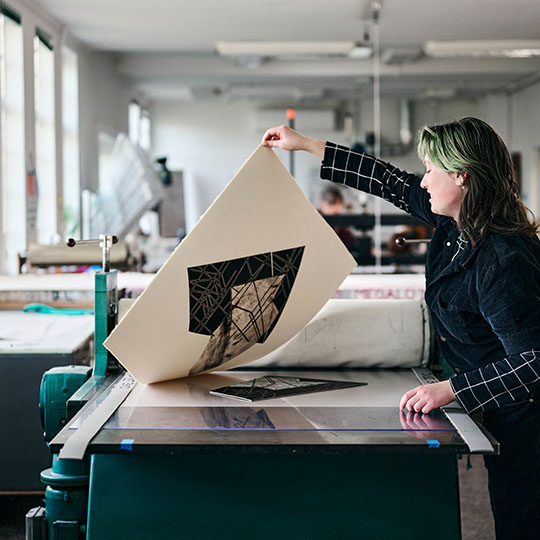 A female artist lifts a print template by one corner from a workstation.