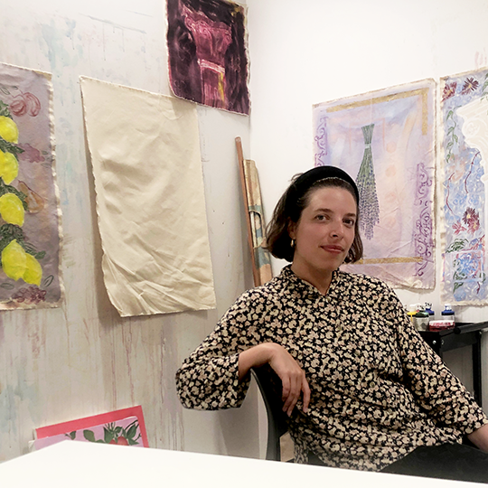 A female artist leans slightly in a chair with several canvas artworks pinned to a wall behind her.