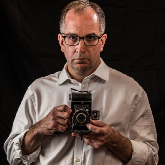Self portrait of David Field, man wearing light tan shirt with cuffs rolled up and dark glasses  looks into camera lens with serious expression on face, he holds a vintage style camera which also appears in the shot.  Black background and warm tone on lens.