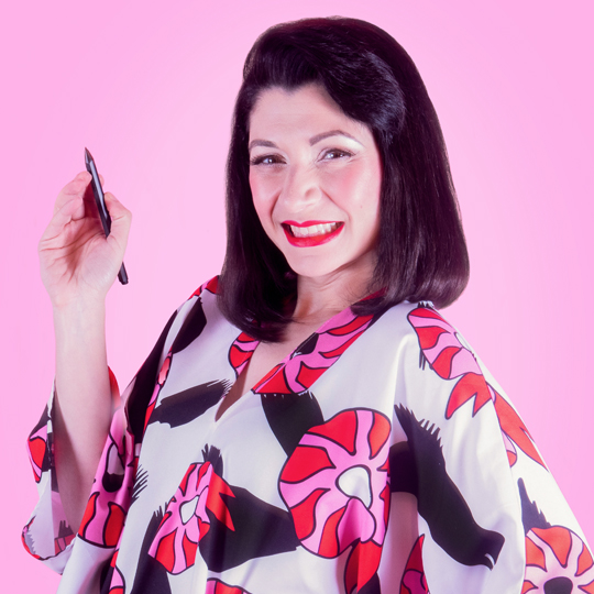 Photo of Stavroula Adameitis, head and shoulder shot, Stavroula wears a pink, black and white shirt, she smiles cheekily at the camera and in one hand she holds a a black pencil . She has dark brown hair to her shoulders with a curled fringe and is wearing pink lipstick.