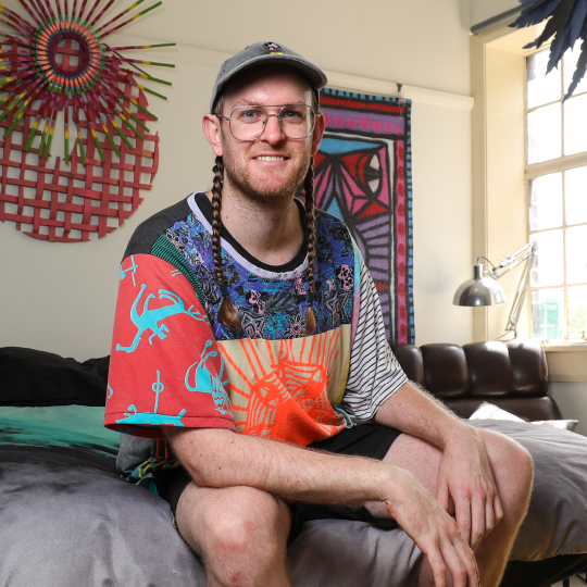 Photo of artist Jeff McCann sitting on bed, cap, hair in two plaits, glasses, printed colourful tshirt, background woven wood artwork, wall hanging, smiling face 