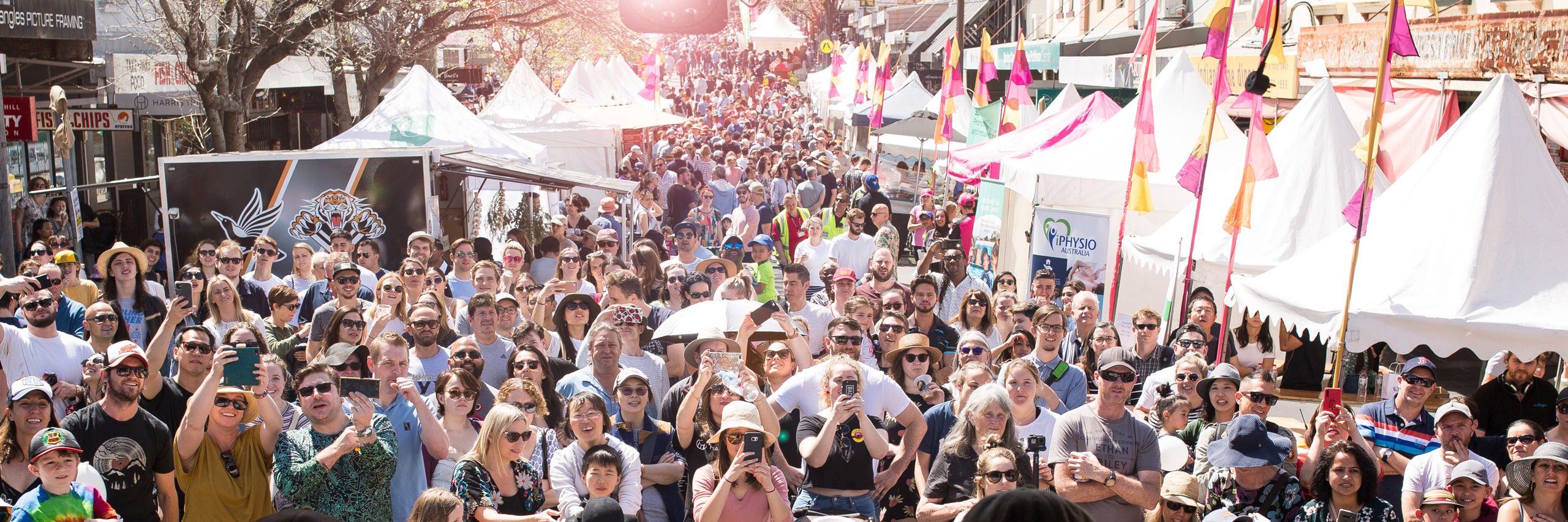 wide shot of a large crowd outside on a sunny day. Mare wearing sunglasses and are holding up mobile phones taking photos