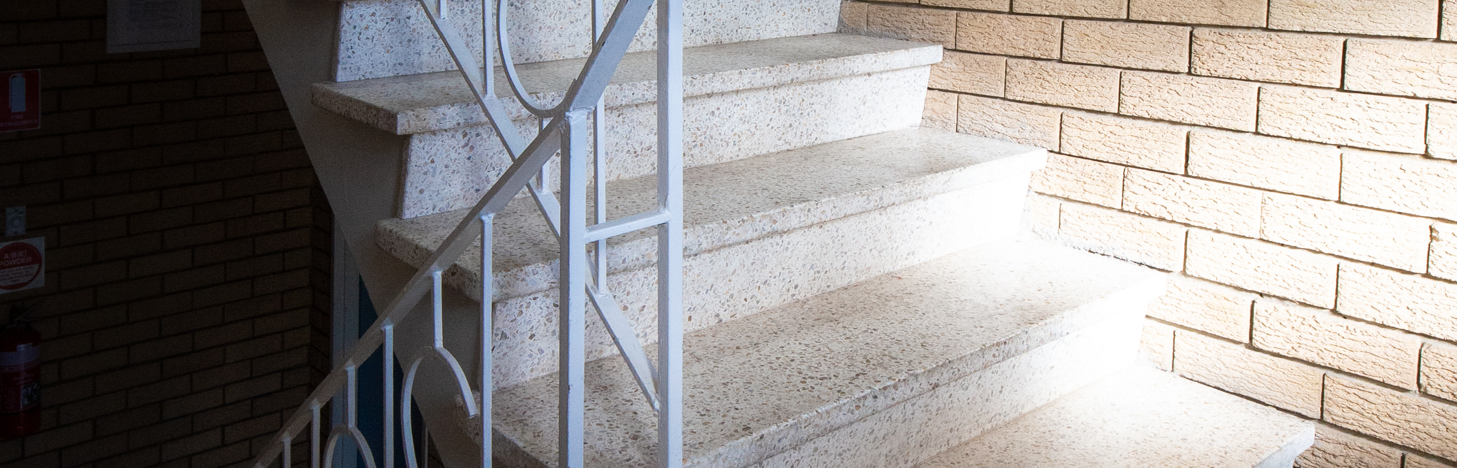 A terraza-surfaced staircase in a walk-up apartment block, bathed in bright sunlight from a window out of view.