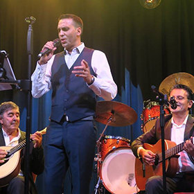 Two men sit down with guitars whilst a man stands and sings