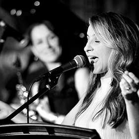 A woman sings into the microphone whilst someone looks on in the background