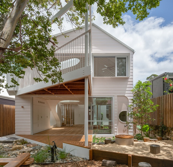 The white cladded rear of a two-storey house with a triangular balcony jutting out into the landscaped yard.