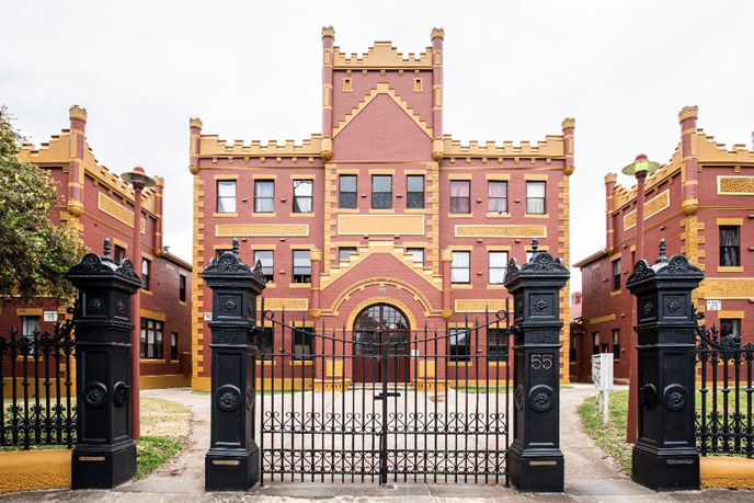 A crenellated earth-coloured building with yellow details behind an imposing black front gate.