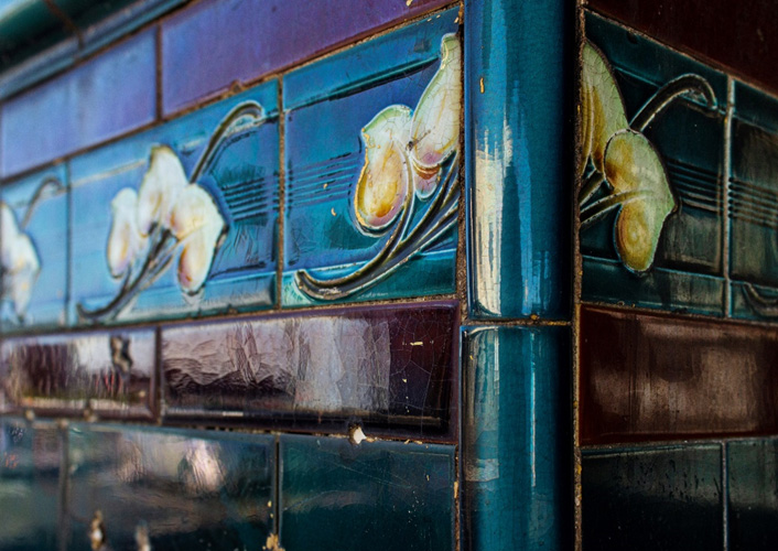 An ornate corner of a building with dark coloured Art Nouveau tiles. Passing car traffic is reflected in the gloss of the tiles.