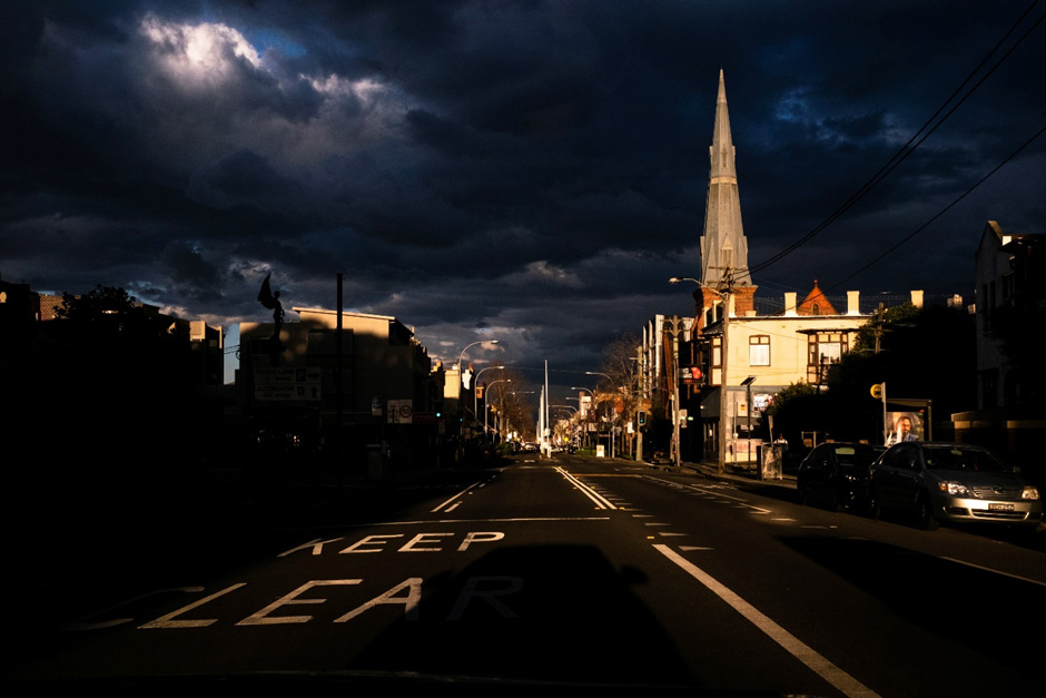 A gloomy view  of an urban main street looking down the roadway. The stormy sky is punctuated with bright sunshine on the shop and church buildings on the right.