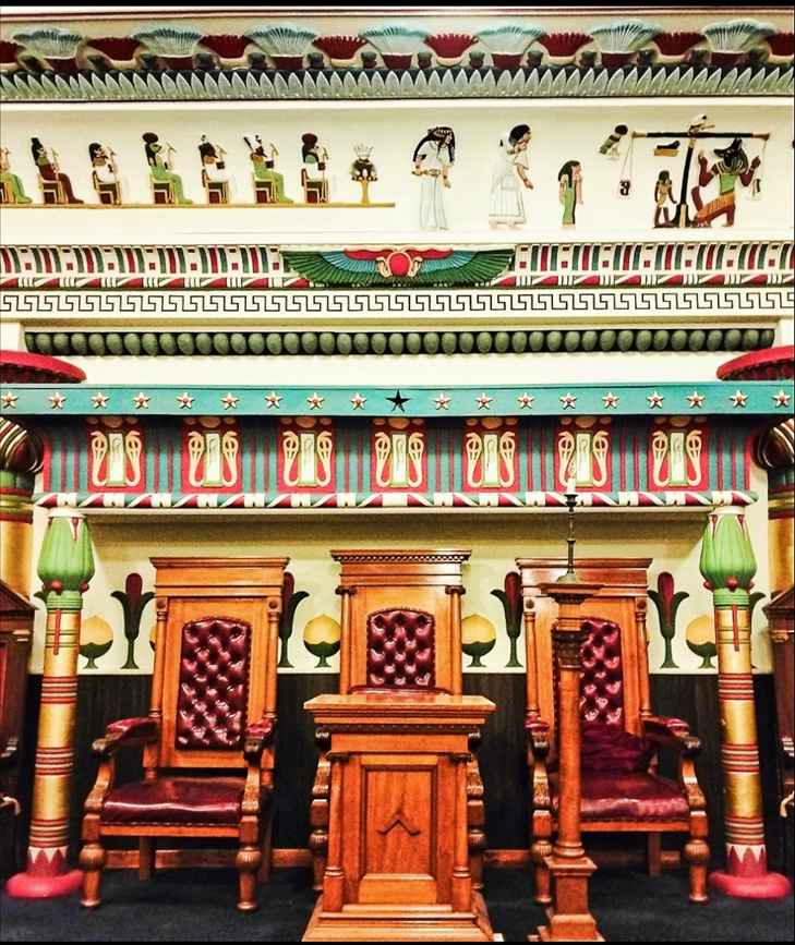 An ornate room with wooden upholstered throne-like chairs. The wall behind is decorated with ancient Egyptian patterns and other ornamentation.