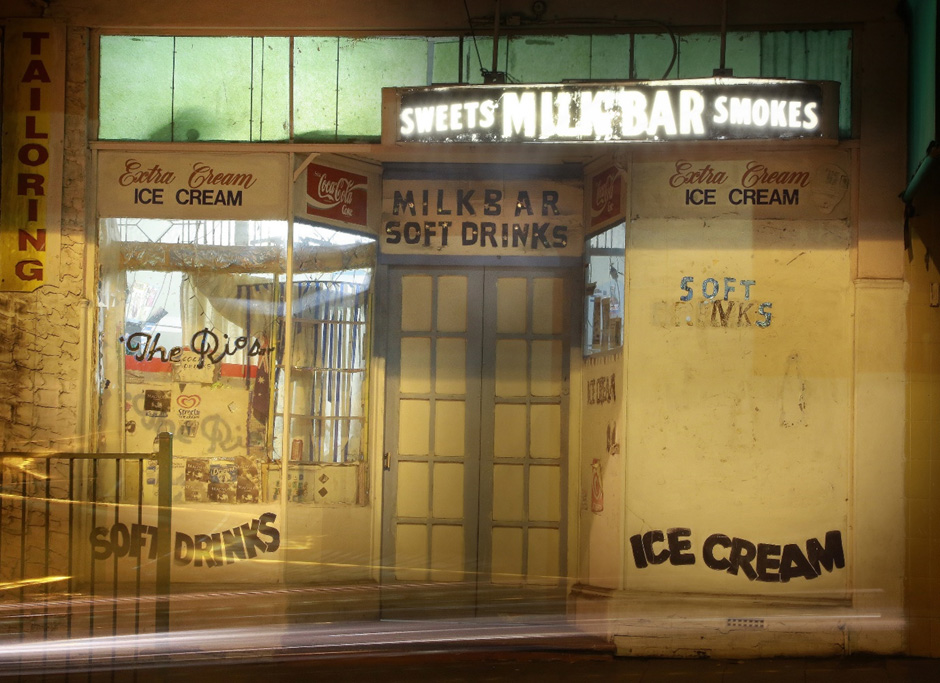 A boarded-up milk bar from a bygone area at night. Despite being closed to the public the interior is nonetheless illuminated.