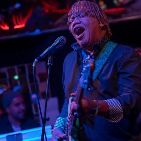 Guitarist and vocalist wearing a navy suit, singing passionately into a microphone, surrounded by blue and red lighting