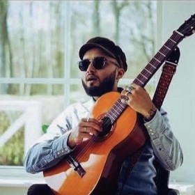 Guitarist sitting down in a white room wearing a white shirt, black cap and circular sunglasses with a guitar propped up on their knee