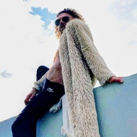 Looking up at a person with beach blonde hair sitting atop a aqua coloured wall, wearing a long fluffy cream jacket, dark jeans and large dark sunglasses