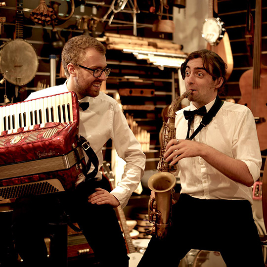 Two smiling musicians - an accordion player and a saxophonist - wearing long-sleeve white shirts and bowties and playing in front of a room of eclectic instruments.