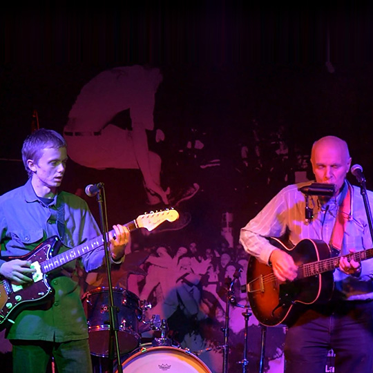 An electric guitarist playing on a stage alongside an acoustic guitarist with an electric harmonica.