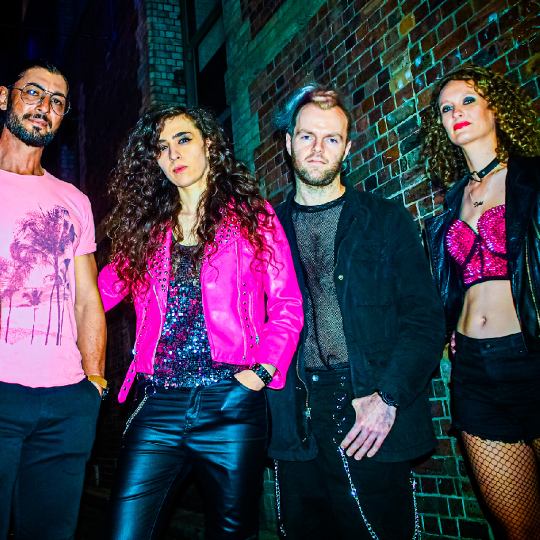 Four people in black and pink coloured rock clothing stand with attitude against a brick wall