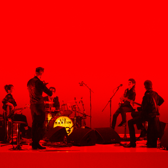 A stylised red and black image of a five-piece rock band playing on stage with their backs turned away from the camera a The band are photographed in black and white tones against a bright red background; the front of the drumkit is bright yellow with text in capital letters that says: Liz Martin