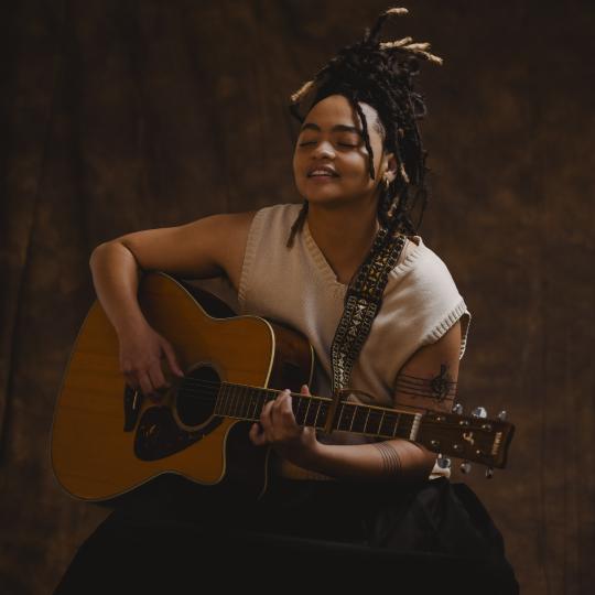 A woman with dredlocks piled high on her head soulfully plays an acoustic guitar with her eyes closed and a half smile