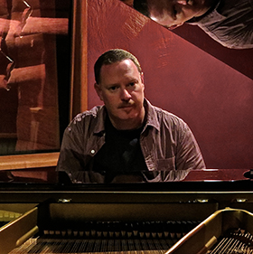 A man sitting at a piano wearing a blue shirt
