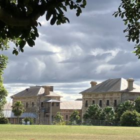 A large building with a lawn in front of it. It showcases outdoor scenery with clouds in the sky, grass, and trees surrounding the building.