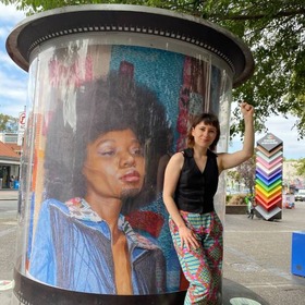 Person with their hand in the air in front of circular with a large photo of a drag king wrapped around it. A rainbow pride coloured beacon is is the background it