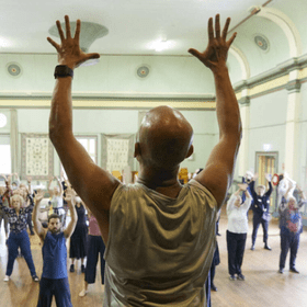 Seniors at a dance class- a bald man in the foreground raises his hands into the air, the crowd facing in copies his movement