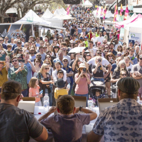 People at a long table eating pizza in from of a large crowd at an outdoor festival