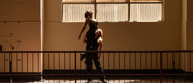 One performer lifts another in an acrobatic pose in a dramatic scene on an industrial stairway landing