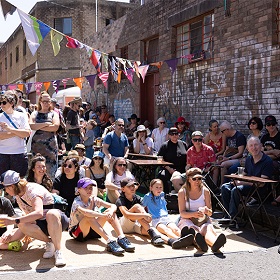 Photo of a crown in a street in St Peters the crowd is sitting on a mat on the ground, sitting in chairs and standing, there is a large building with raw brick behind them with white graffiti on it, bunting is strung up making a triangle shape above their heads