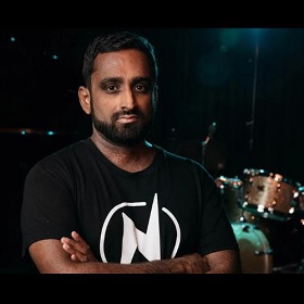 A man looks straight at the camera with arms folded, he has brown skin and dark facial hair, he has a serious expression on his face, behind him is a drum kit