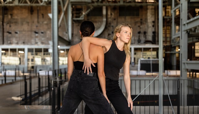 Photo of two Legs On The Wall dancers one has her back to teh camera the others hand fans out on the dancers back and she stands profile with long blond hair, both dancers wear black jeans and singlets