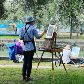 Painter stands outside on the GreenWay painting on an easel, they have a paintbrush in their hand and you can see trees and the canal in the background