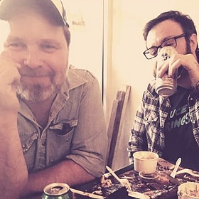 Photo of Adam Young and Sam Shinazzi at a table drinking beer in cans, the table they are sitting at appears to have food on it, Adam wears a cap and smiles at the camera, Sam wears black framed rectangle glasses and holds a beer can up to his face