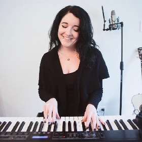 Musician Alicia May sits in studio at keyboard pressing keys, she wears black clothing and smiles with eyes shut, she has long hair. In the background is a white wall and a microphone on a stand