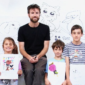 Photo of three kids with a grown up in the middle. the grown up appears to be an art teacher and holds a marker in his hands, he wears a black t-shirt and a beard, the children hold up pieces of paper with their cartoons , in the background are cartoon figures drawn on a large white background, a kookaburra, cat like creature and a bunny.