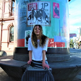 Photo of artist Karen Farmer sitting on the Newtown ArtSeat, she has her hands on her knees, smiling with long curly brown hair covering her face, she wears a white t-shirt and a pleated silvery grey silky long skirt. In the ArtSeat is Karen\